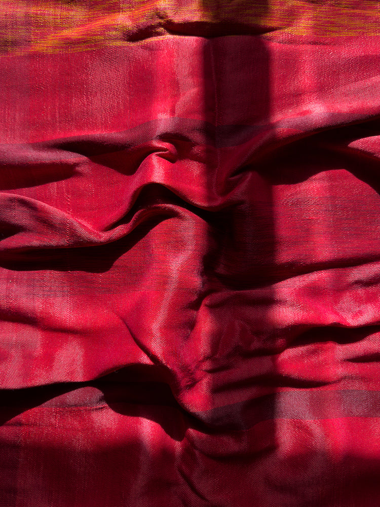 A close-up shot of a textured, red fabric with horizontal stripes of yellow and orange. The fabric is draped and folded, creating shadows and highlights that emphasize its texture and sheen. A shadow of vertical lines falls across the fabric.