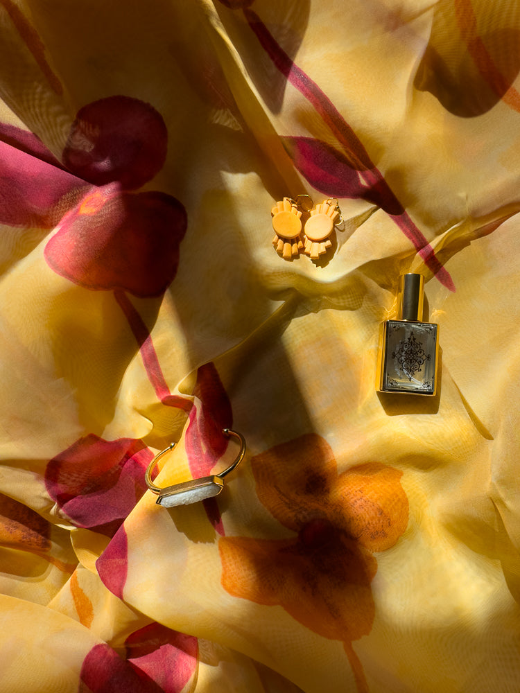 A flat lay of beauty products on a yellow floral scarf. A gold-capped perfume bottle, a gold cuff bracelet with a white stone, and a pair of orange earrings are arranged on the fabric.