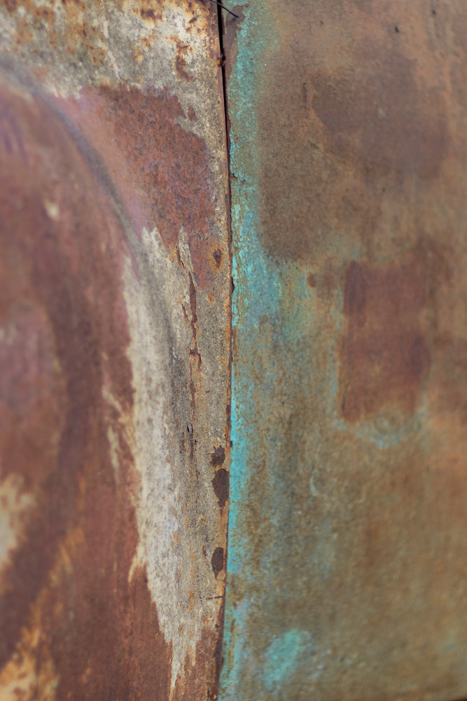 Close-up of weathered, rusted metal with patches of peeling turquoise paint. The surface shows texture and discoloration, with a seam running vertically down the center.