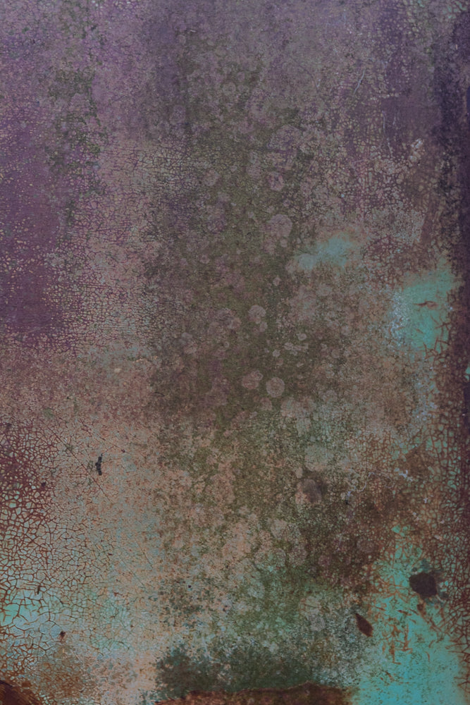 A close-up, abstract view of a weathered surface with peeling paint in shades of purple, green, and brown. The texture is characterized by fine cracks and irregular patches, suggesting age and decay.