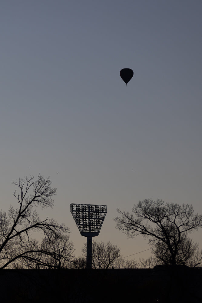A hot air balloon floats in the dusky sky above silhouetted trees and a stadium light tower.