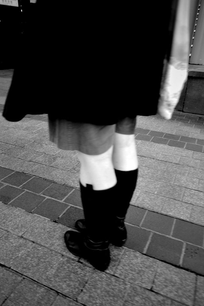 A person wearing a black skirt, white socks, and black boots stands on a tiled sidewalk.