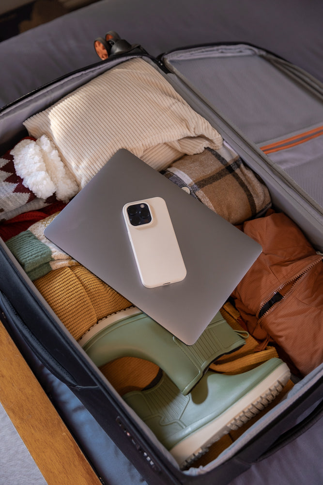 A suitcase is packed with clothes, a laptop, and a smartphone. The contents include a cream-colored sweater, a plaid shirt, a brown jacket, green boots, and other folded garments.