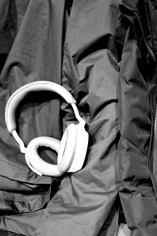 A pair of white over-ear headphones rests on a dark, wrinkled fabric, possibly a jacket or coat. The headphones are positioned diagonally across the frame, with one earcup and part of the headband visible. The fabric has a textured appearance with folds and shadows, creating a dramatic contrast with the smooth, light-colored headphones.