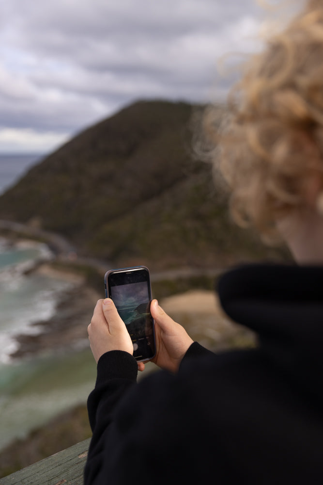A person with curly blonde hair, wearing a black hoodie, holds a smartphone in their hands, capturing a scenic view of a coastline with waves crashing on the shore and a winding road along a hill.