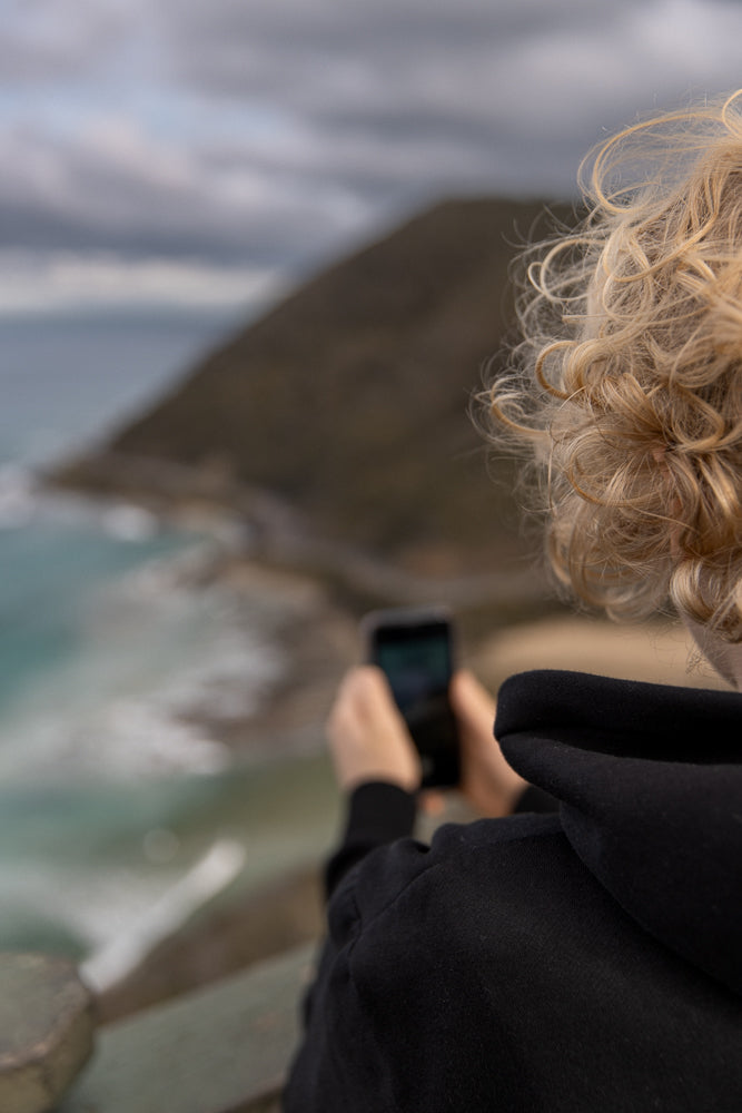 A person with blonde curly hair, wearing a black hoodie, holds a smartphone in their hands, capturing a photo of a coastal landscape with waves and a cloudy sky.