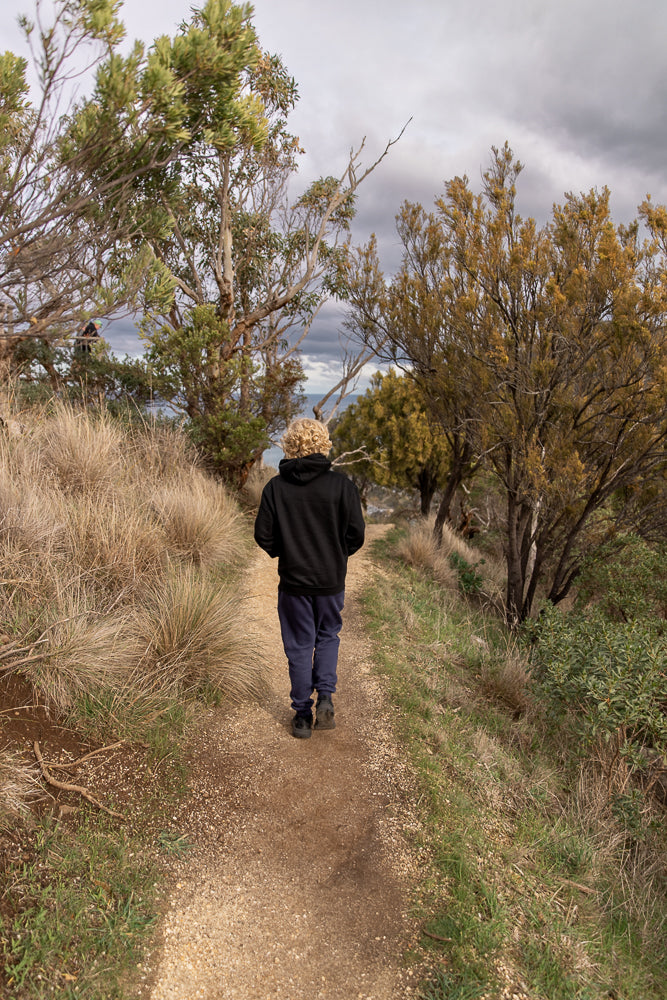A person with curly blonde hair walks away from the camera on a dirt path through a natural landscape with trees and dry grass. The sky is cloudy.