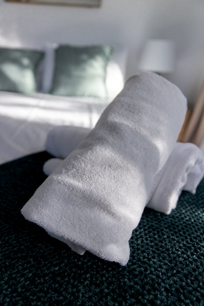 Two neatly rolled white towels are placed on a dark green textured surface. In the background, a bed with white linens and two light green pillows is visible, along with a bedside lamp.