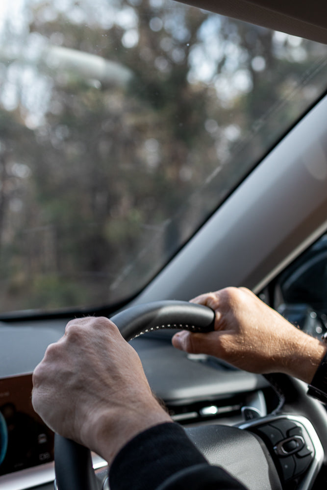 A person's hands grip the steering wheel of a car, with the dashboard and windshield visible in the background. The view is from inside the vehicle, looking forward.