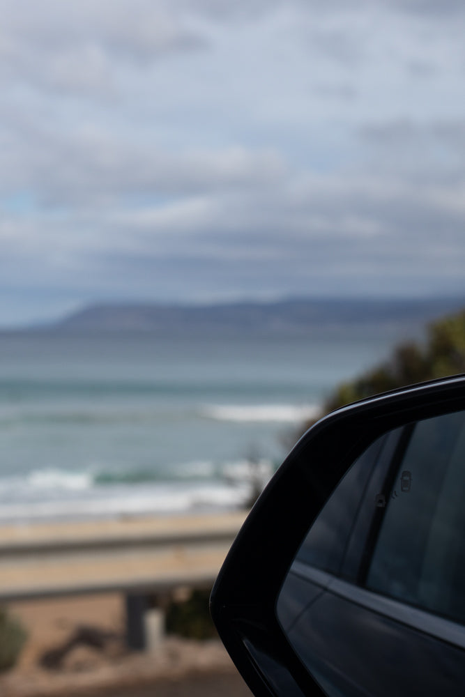 A car's side mirror reflects a coastal landscape with the ocean and distant hills under a cloudy sky.