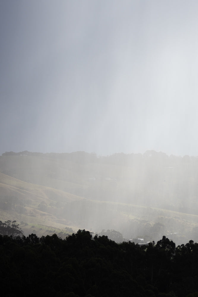 Rain falls on rolling silhouette hills and trees under a cloudy sky with a touch of blue