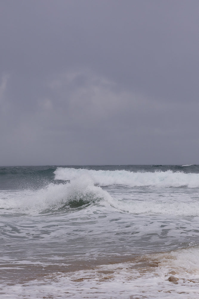 A grey, overcast sky hangs over a choppy ocean with several waves breaking. White foam and spray are visible as the waves crash onto the shore.