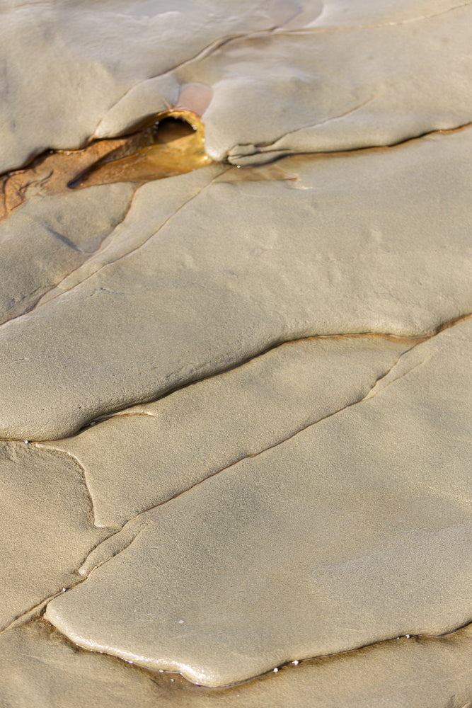 Close-up of wet sand with natural lines and indentations. A small pool of water reflects the light in the upper left corner.