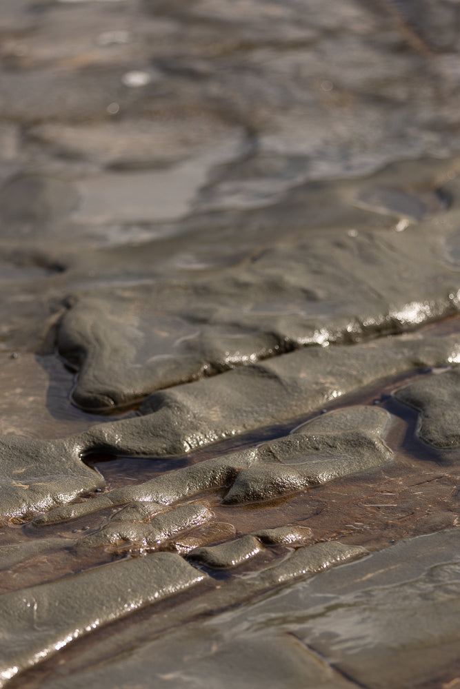 Close-up of wet, textured sand and rock formations with shallow pools of water reflecting sunlight. The patterns create a natural, abstract landscape.