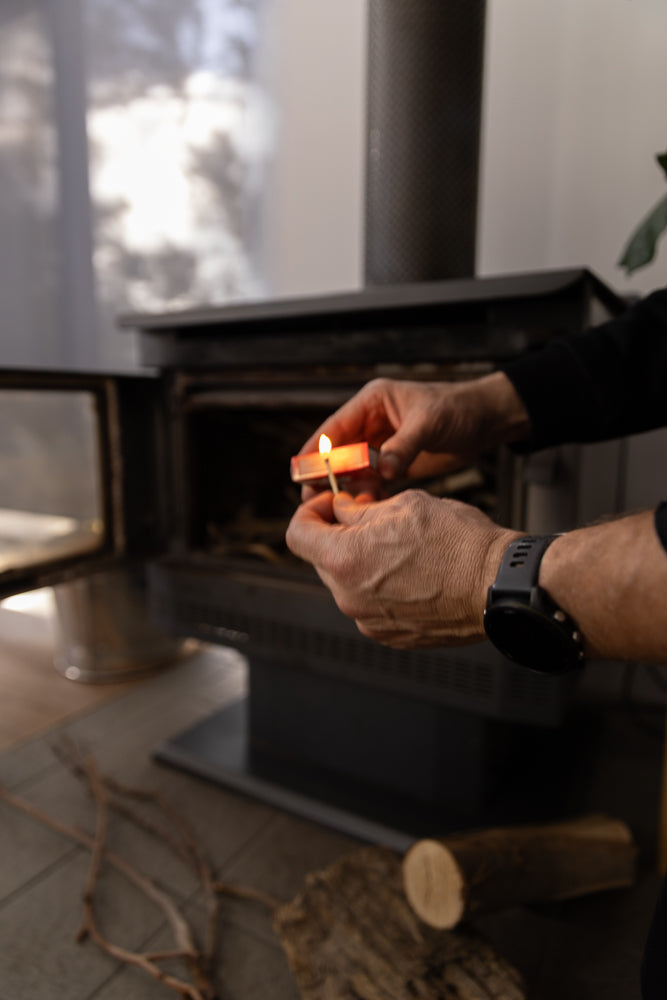 A person lights a match to start a fire in a wood-burning stove. The stove has an open door, and logs are visible on the hearth.