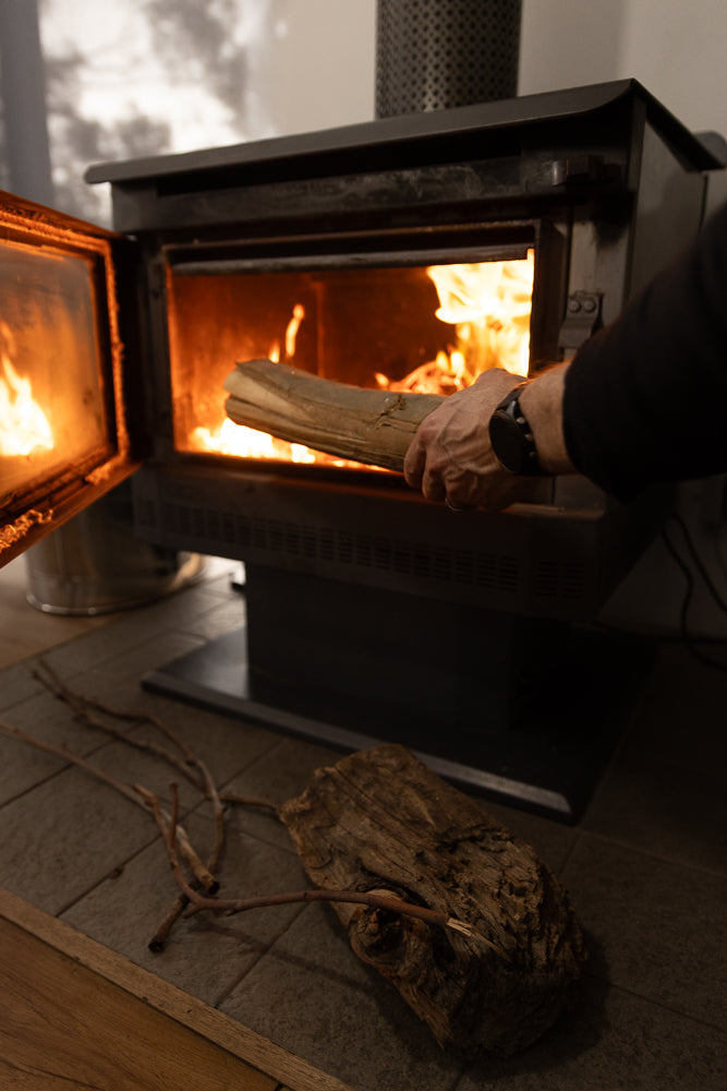 A person's hand places a log into a modern wood-burning fireplace with an open glass door. Flames are visible inside the firebox, and a few pieces of wood and twigs are on the floor in front of the fireplace.