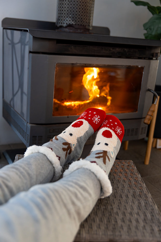 A person's legs wearing fuzzy grey pants and festive Christmas socks with reindeer and snowflakes are resting in front of a wood-burning fireplace.