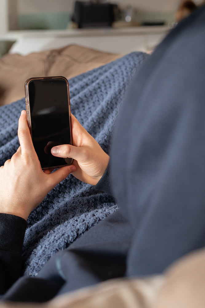 A person is holding a smartphone in their hands, with their thumbs resting on the screen. They are wearing a dark blue hoodie and are covered by a blue knitted blanket.