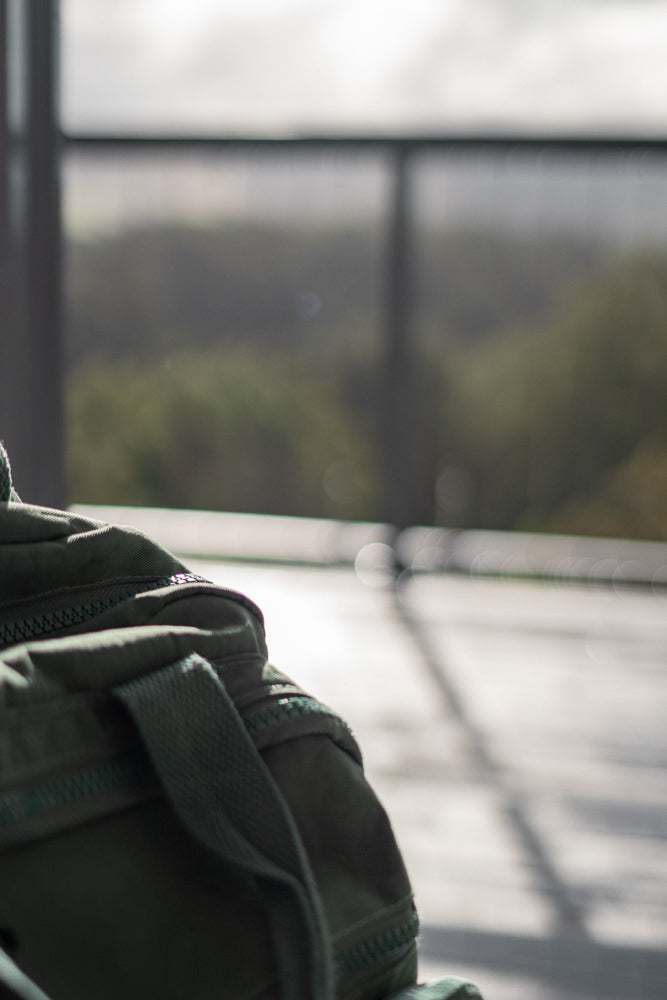 A close-up shot of a green canvas duffel bag with a zipper and strap visible. The background is blurred, showing a hint of trees and a railing.