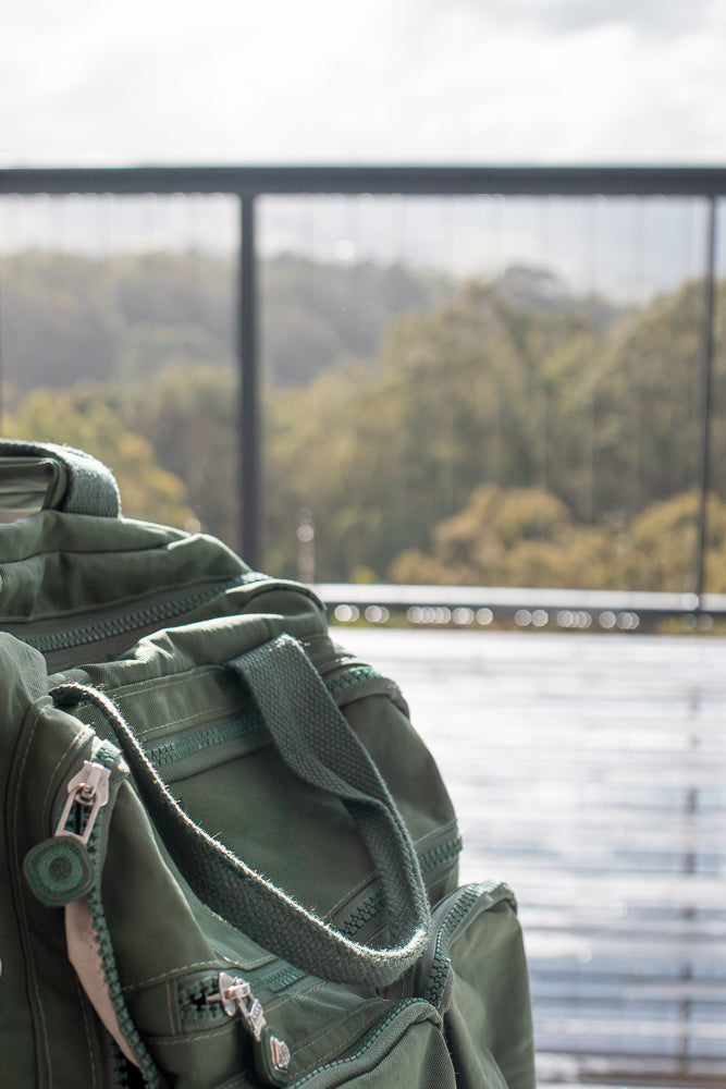 A close-up of a green duffel bag with zippers and straps. The bag is placed on a wooden surface, with a blurred background of trees and a railing.