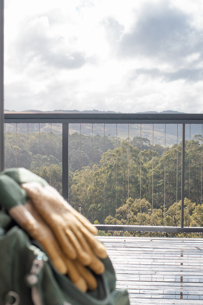 A pair of tan work gloves rests on a green bag on a wooden deck. Beyond the deck, a railing separates the view of a lush green forest and rolling hills under a cloudy sky.