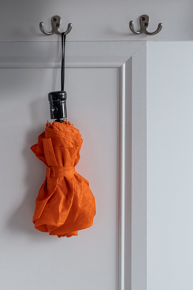 A closed, bright orange umbrella hangs from a double hook mounted on a white wall. The umbrella is tied with a fabric strap and has a black handle with a silver button.