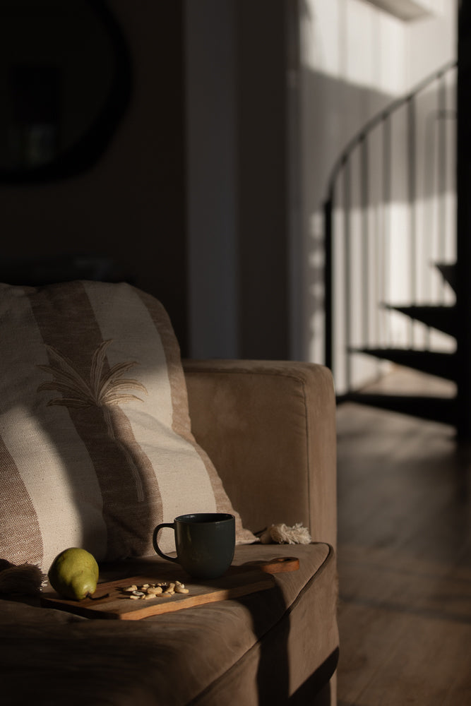 A mug of coffee sits on a wooden board with a pear and nuts on a sofa next to a striped pillow. Sunlight streams in from the right, casting shadows.