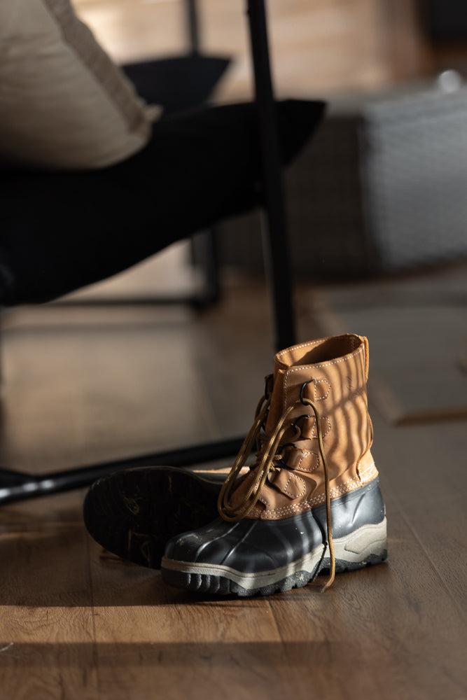 A pair of tan and black waterproof boots with laces are on a wooden floor. The boots are in the foreground, with a chair and pillow blurred in the background.