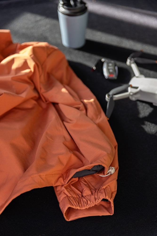 An orange jacket is laid out on a dark surface next to a light blue travel mug and a white drone. The jacket is wrinkled and the sleeve is pulled back, revealing a dark lining and a drawstring.