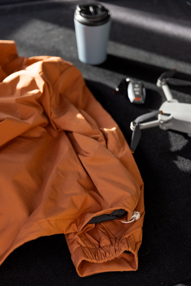 A close-up shot shows a crumpled orange jacket with a drawstring cuff. In the background, a light blue travel mug and a white drone with black propellers are visible on a dark surface.