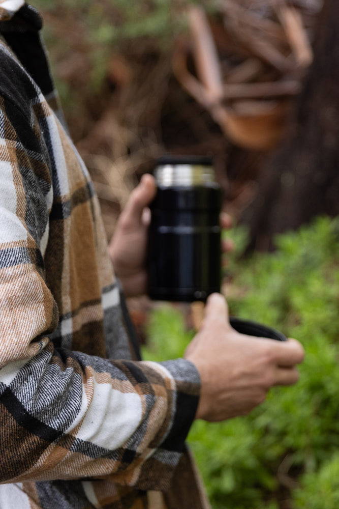 A person wearing a plaid shirt holds a black thermos and a cup outdoors with green foliage in the background.