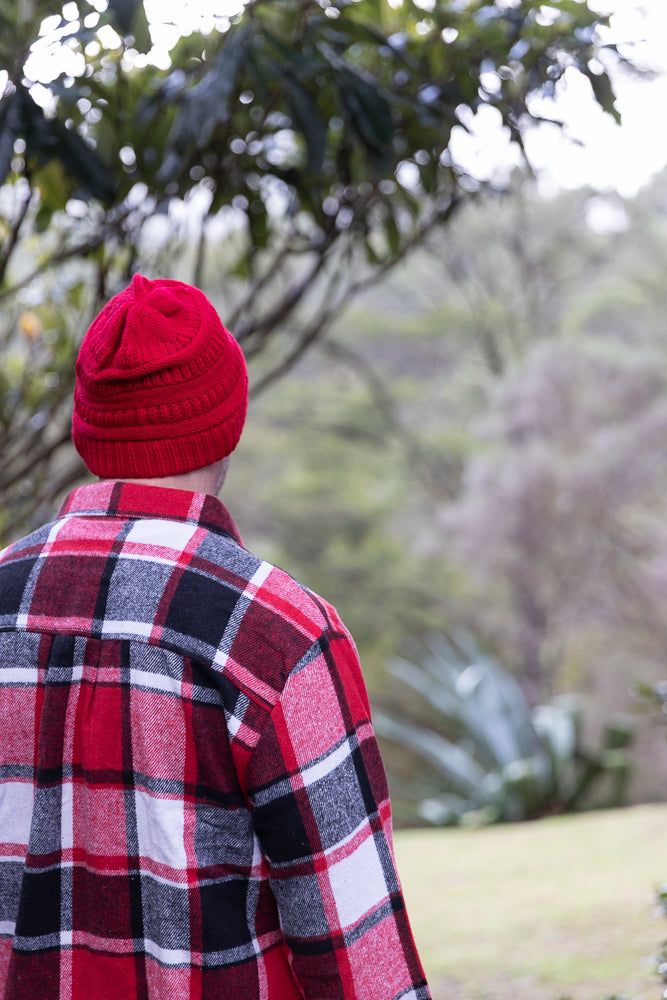 A person wearing a red knitted beanie and a red, black, and white plaid shirt stands with their back to the camera, looking out at a blurred natural landscape.