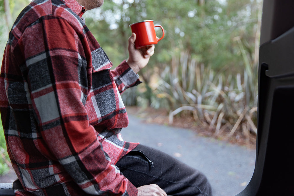 A person wearing a red and black plaid shirt sits in the back of a vehicle, holding a red enamel mug. The background is blurred with greenery and trees.
