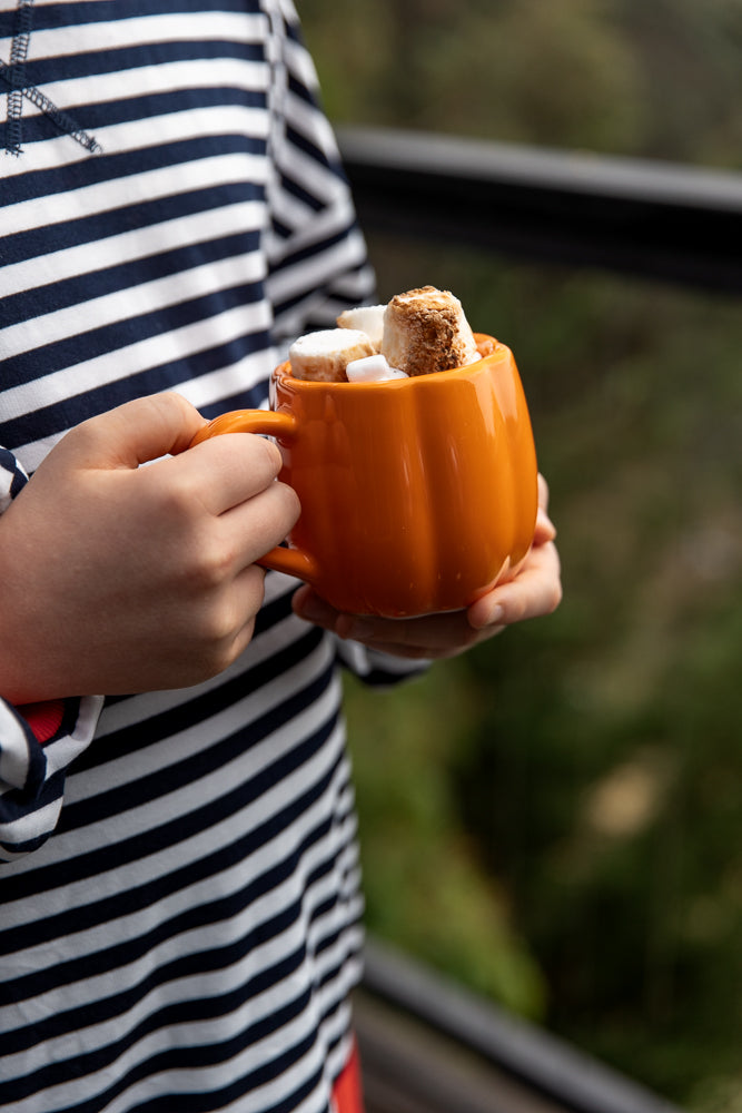 A person wearing a navy and white striped shirt holds an orange pumpkin-shaped mug filled with hot chocolate and marshmallows.