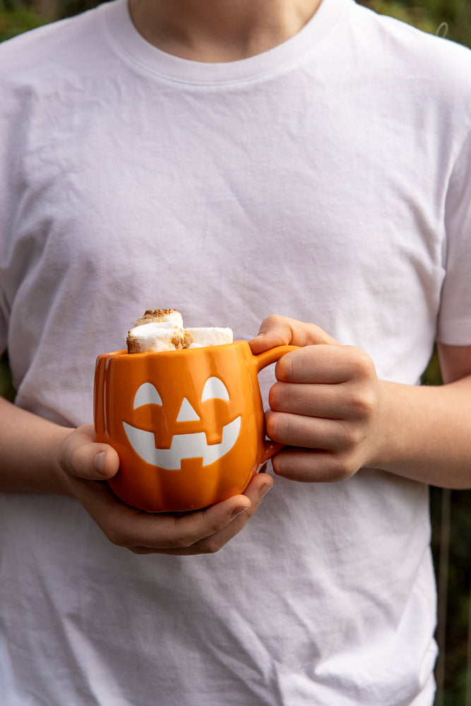 A person in a white t-shirt holds a pumpkin-shaped mug filled with marshmallows. The mug has a carved jack-o'-lantern face.