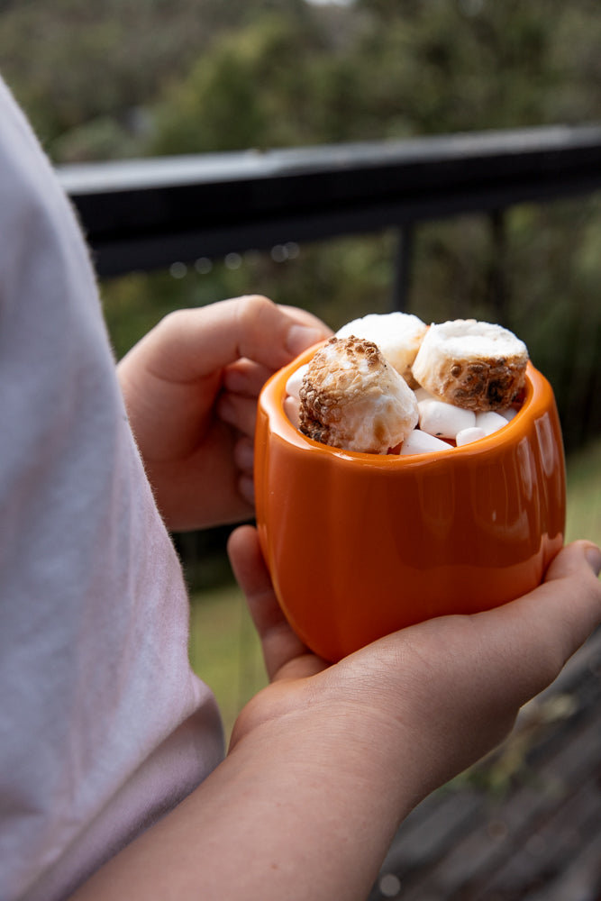 A person holds an orange mug filled with hot chocolate and marshmallows, enjoying a warm drink outdoors.