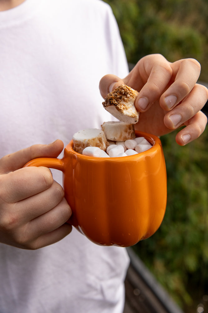 A person holds an orange pumpkin-shaped mug filled with marshmallows and a toasted marshmallow being added to the top.