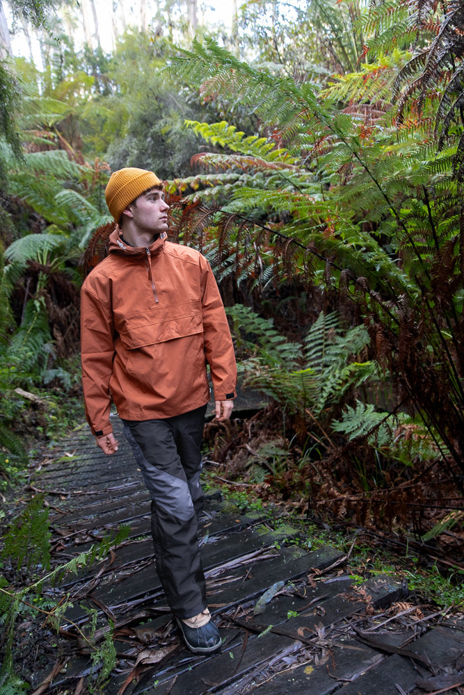 A young man wearing an orange beanie and jacket walks on a wooden path through a lush forest with ferns.