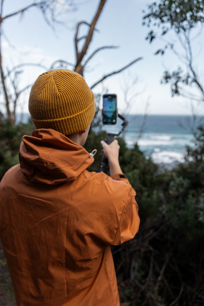 A person wearing an orange beanie and jacket films the ocean with a smartphone on a gimbal.