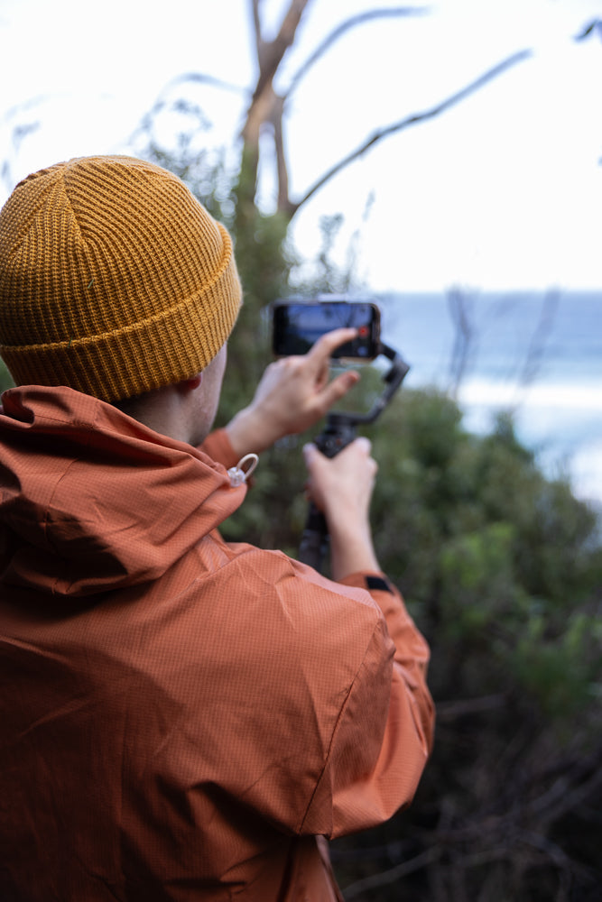 A person wearing an orange jacket and a yellow beanie films the ocean with a smartphone mounted on a gimbal.