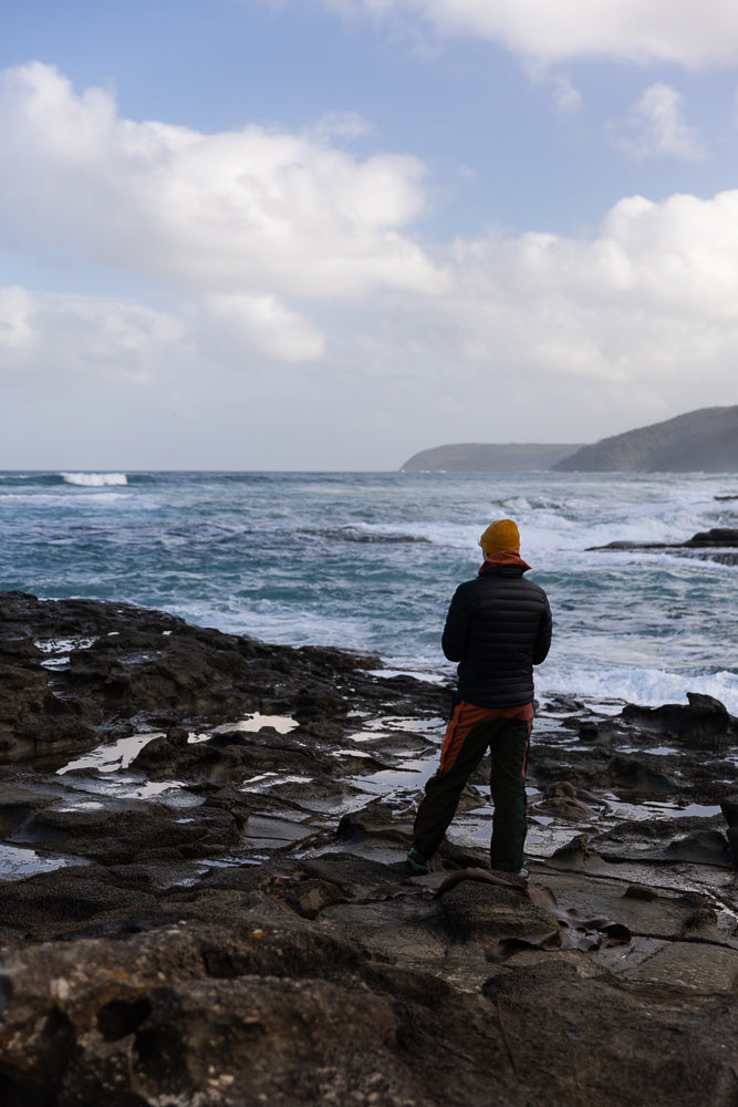 A person wearing a yellow beanie and a dark puffer jacket stands on dark, wet rocks, looking out at the choppy blue ocean under a cloudy sky.