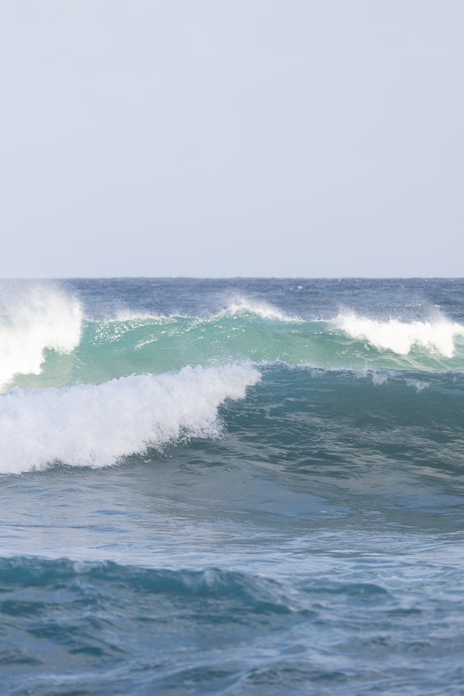 A series of ocean waves roll towards the shore under a pale blue sky. The waves are a mix of deep blue and turquoise, with white foam cresting on the larger waves.