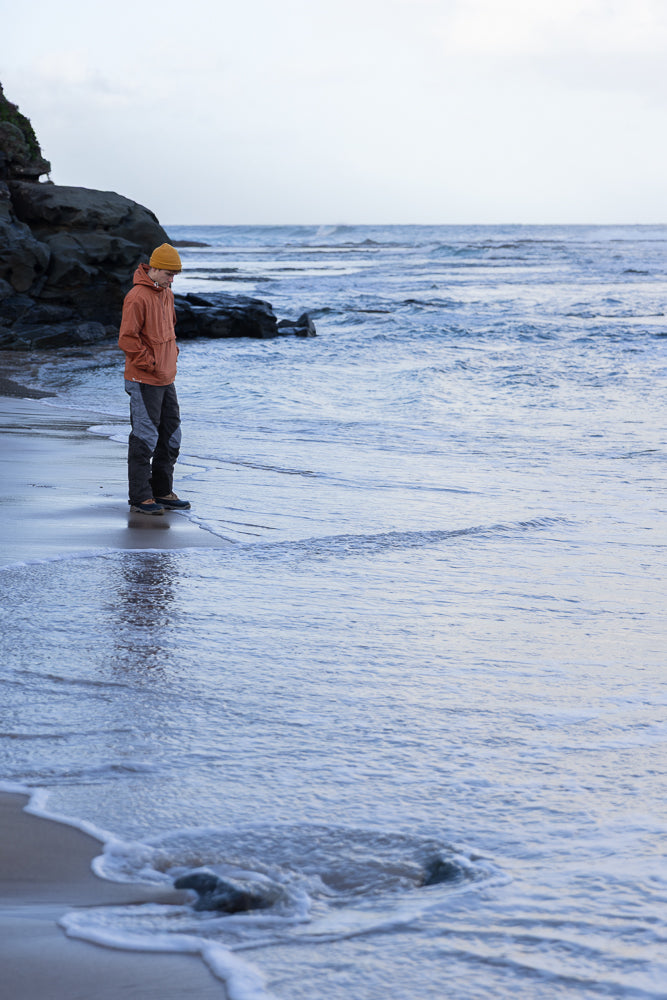 A young man wearing an orange beanie and jacket stands on a sandy beach, looking down at the incoming tide. Dark rocks and vegetation are visible on the left side of the frame, with the ocean stretching out to the horizon under a cloudy sky.