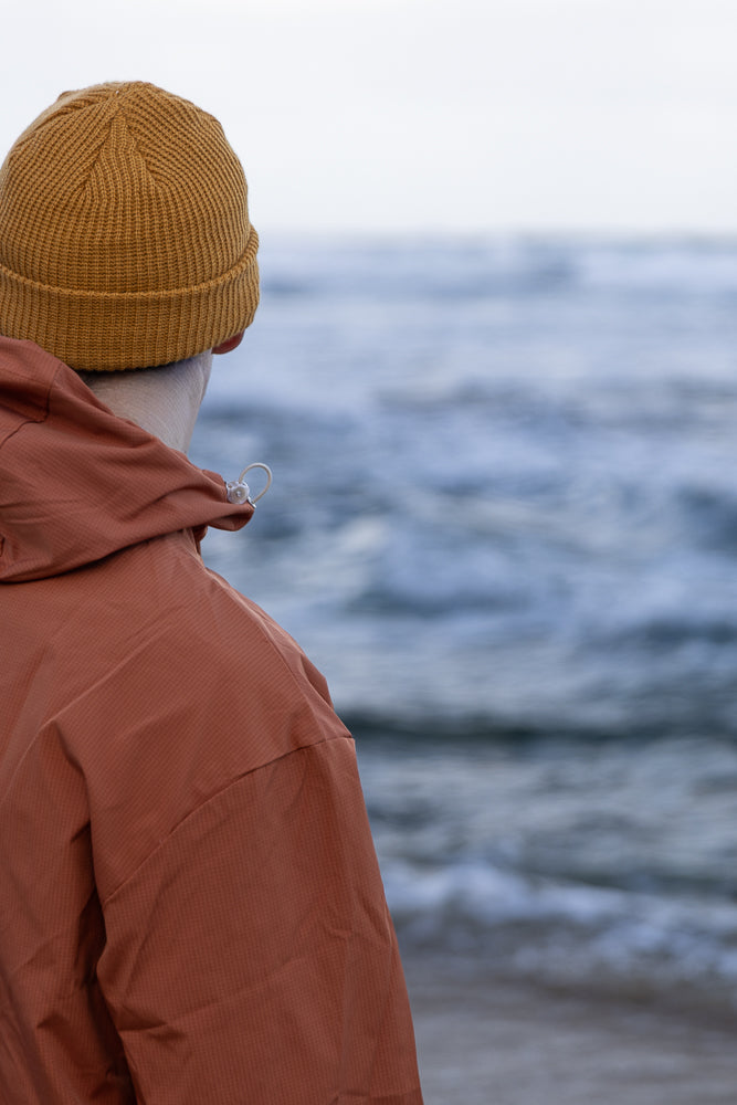 A person wearing a rust-colored jacket and a mustard-yellow beanie looks out at the ocean. The water is choppy with whitecaps, and the sky is overcast.