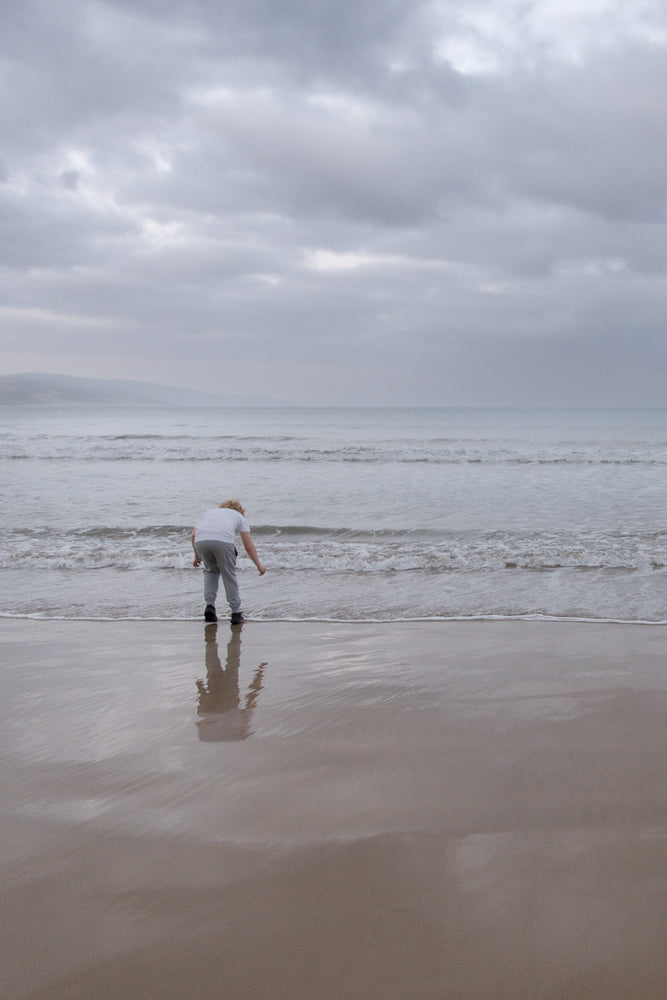 A child with blond hair wearing a white t-shirt and grey pants bends down to touch the water at the edge of the sea on a cloudy day. The wet sand reflects the child and the sky.