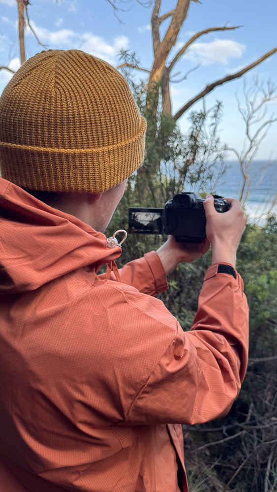 A person wearing a mustard yellow beanie and an orange jacket films the ocean with a black camera. The background shows trees and a blue sky with clouds.