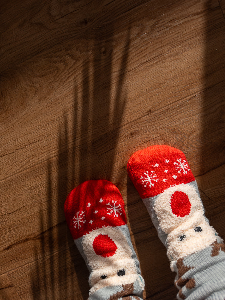 A pair of feet wearing cozy, festive socks with a reindeer and snowflake pattern, resting on a wooden floor. Sunlight casts shadows across the floor.