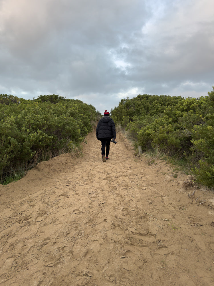 A person wearing a black puffer jacket and a red and white beanie walks away from the camera down a sandy path lined with green bushes under a cloudy sky. The person is holding a camera in their right hand.
