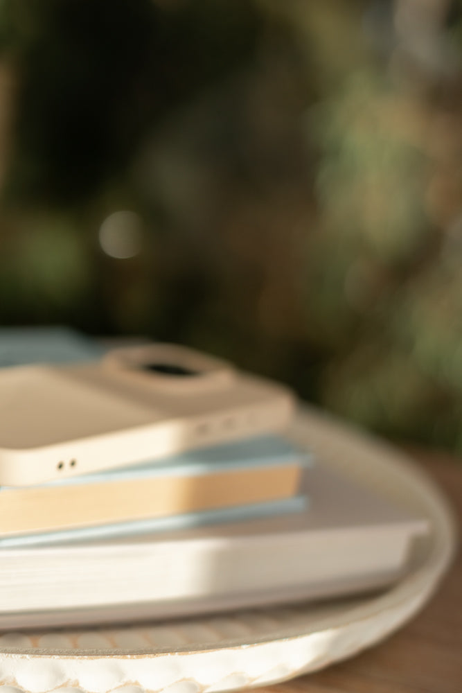 A close-up, slightly blurred shot shows a stack of books on a decorative tray. A light-colored phone case rests on top of the books, with the camera lens visible.