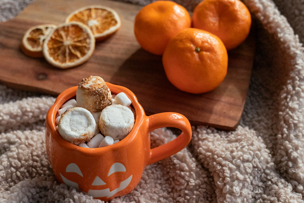 A Halloween-themed orange mug shaped like a jack-o'-lantern is filled with marshmallows and topped with toasted marshmallows. In the background, three clementines sit on a wooden board next to dried orange slices.